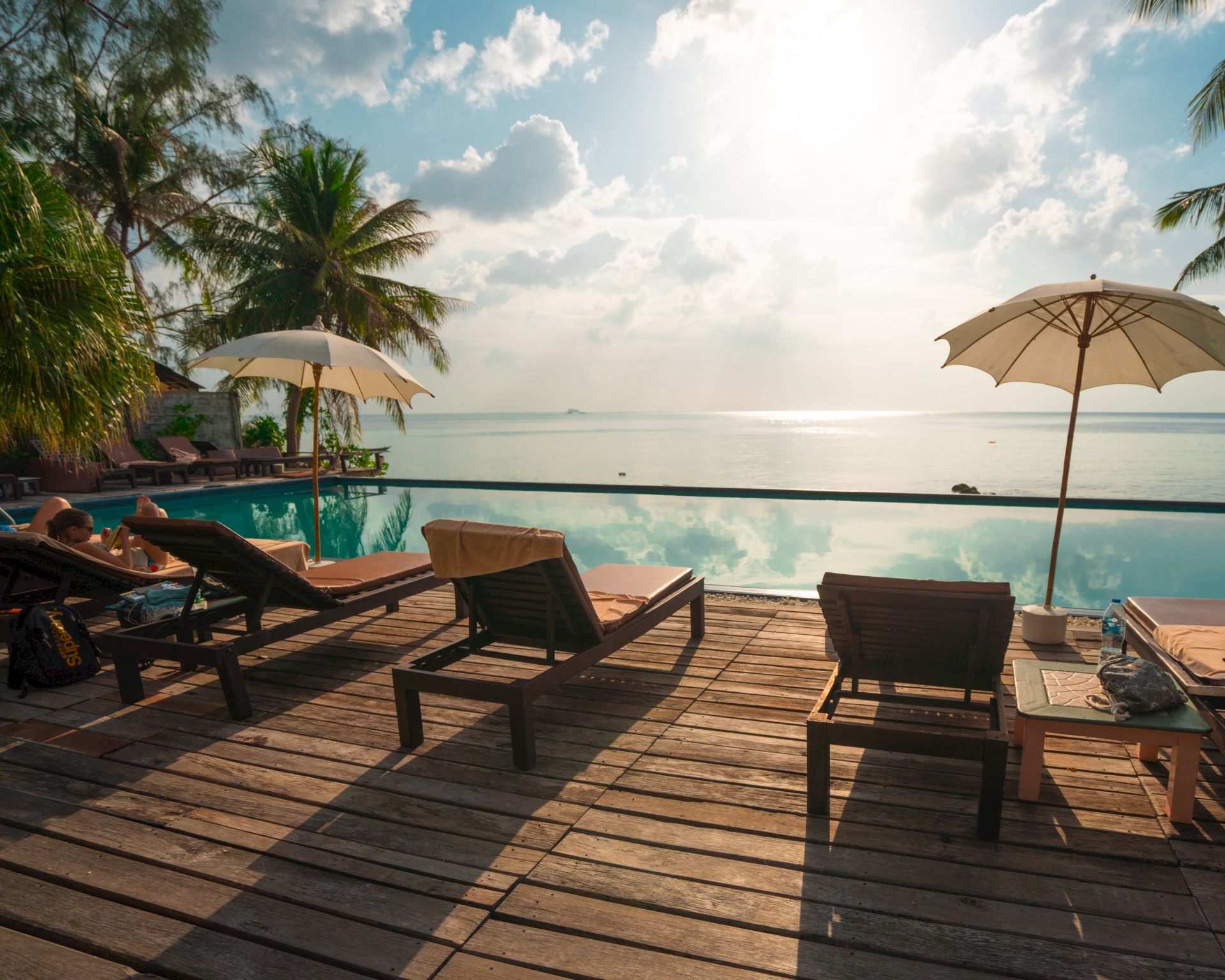 A tropical beachfront resort with lounge chairs, two umbrellas, a swimming pool, palm trees, and a bright ocean under blue skies.