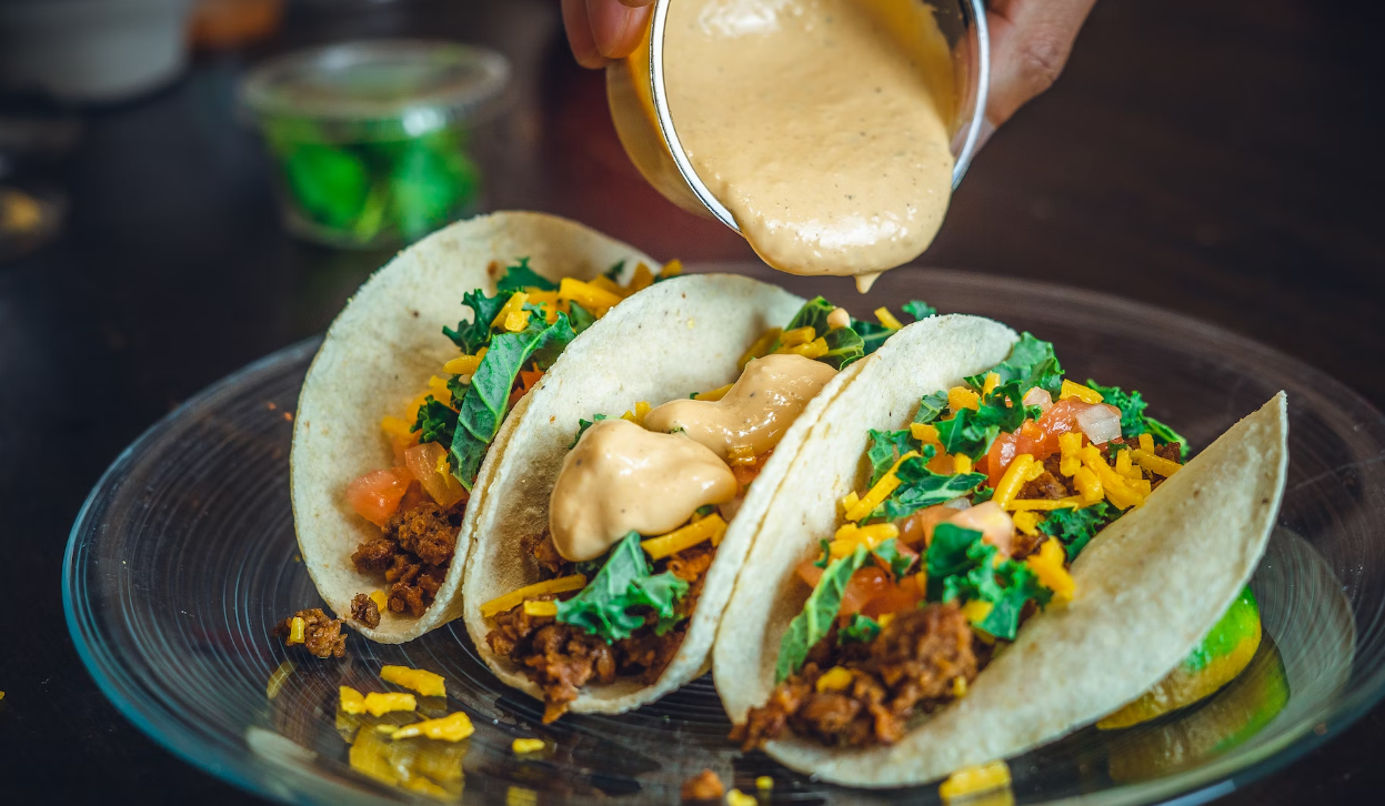 Three tacos with shredded meat, cheese and veggies on a plate, with creamy sauce being poured over them.