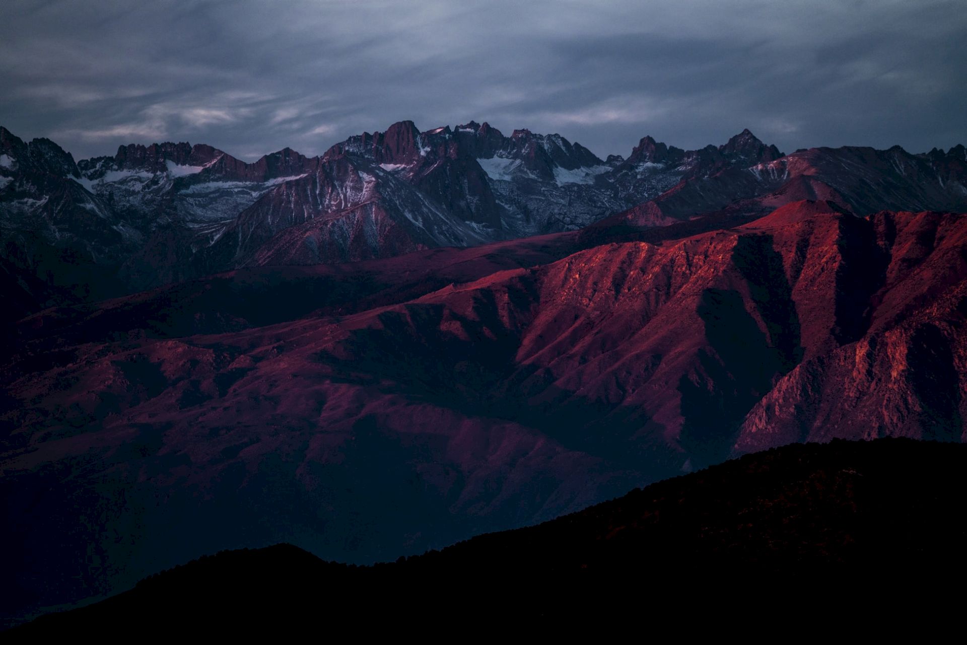 Snowy mountain range at dusk with rugged ridges glowing red-purple under stormy clouds.