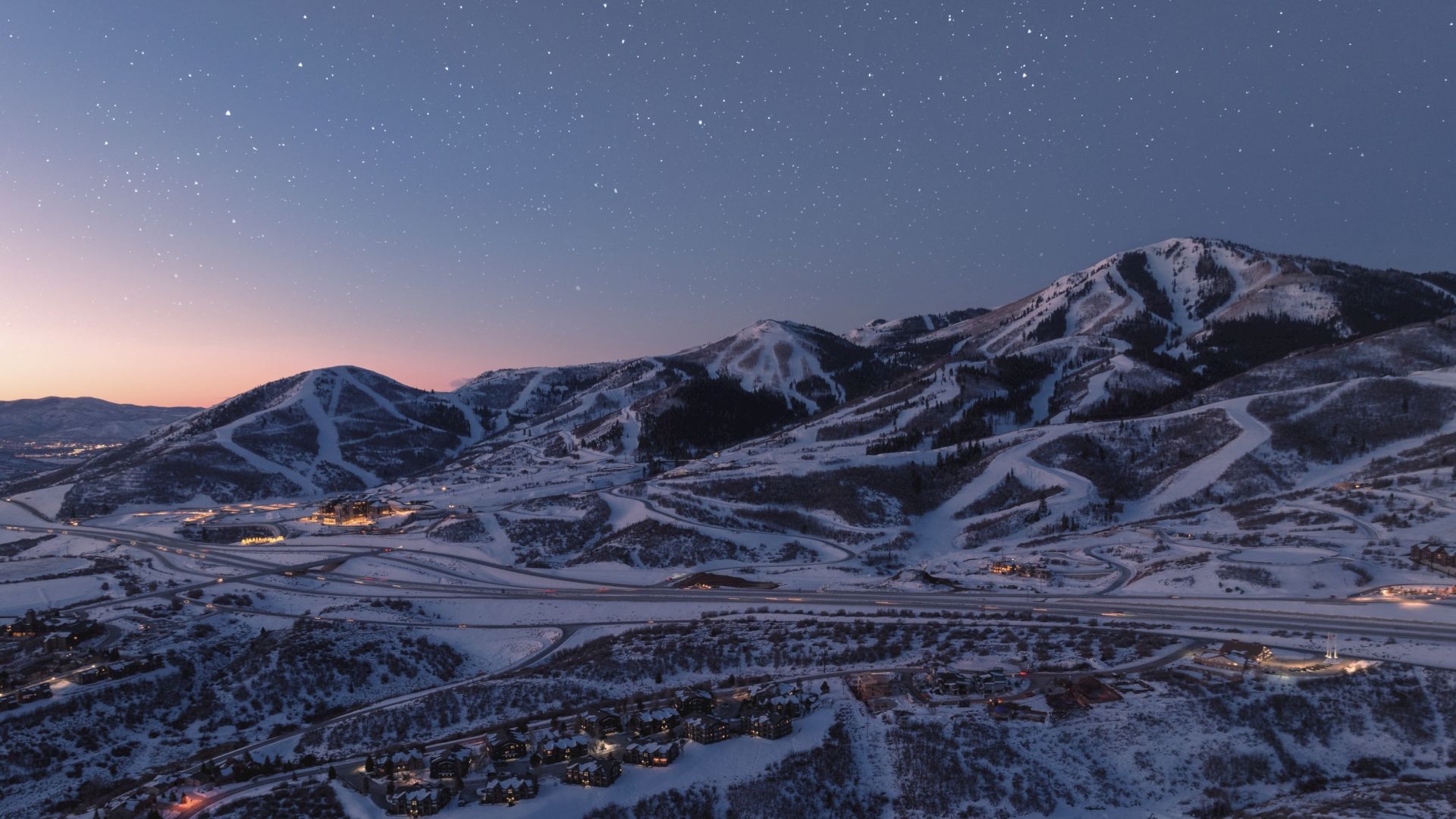 A snowy mountain landscape at dusk with starry sky, illuminated roads and a small cluster of buildings in the valley below.