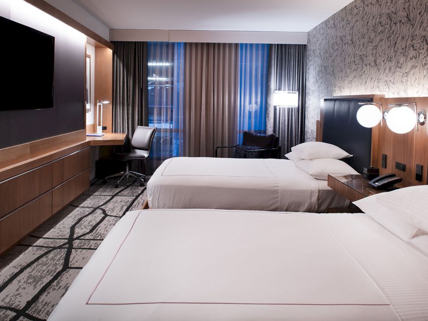 A modern hotel room with two neatly made beds, dark headboards, and curtains beside large windows.