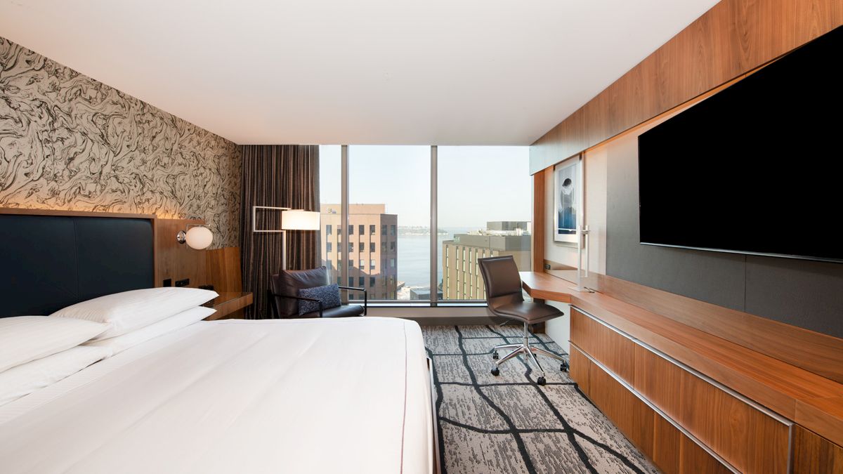 A modern hotel room: large bed, desk chair, two wall-mounted TVs, and windows showing city buildings.