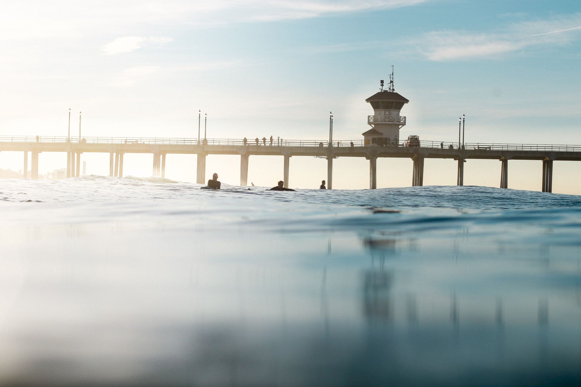 A calm harbor scene: swimmers in the water beneath a pier with a lifeguard tower at sunrise.